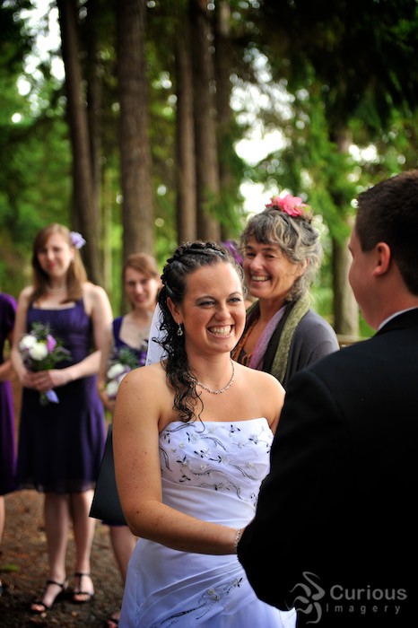 ecstatic bride smiles at groom during outdoor wedding ceremony. female officiant in background.